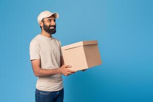 A smiling bearded Indian delivery man in a light cap and beige t-shirt holds a brown box in front of a bright blue background, copy space photo