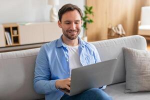 A man is sitting on a couch while using a laptop. He appears focused on the screen, typing and clicking. The room is well-lit, with a coffee table in front of him. photo