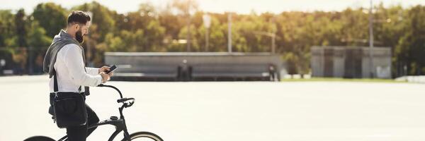 Modern businessman checking email on phone, while riding bicycle by park to work, panorama with copy space photo