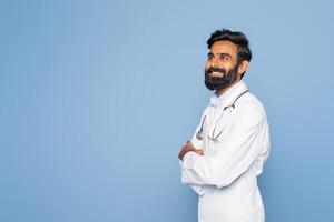 A bearded Indian doctor wearing a white coat and stethoscope smiles while standing confidently against a blue studio background. His arms are crossed, conveying professionalism and warmth. photo