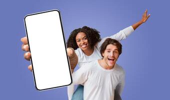 A young multiracial couple is holding a smartphone with a blank screen in front of them. The woman is giving a thumbs up while riding on the man's back. photo