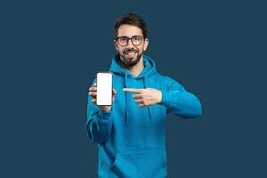 A young man stands against a solid blue background, wearing a blue hoodie and glasses. He is pointing at his smartphone with a cheerful expression, seemingly excited about what he is sharing. photo