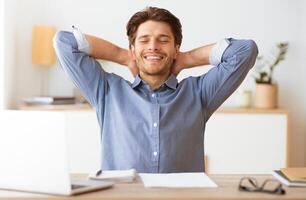 A relaxed man sits at his desk, leaning back with his arms behind his head, smiling and taking a break from work. He appears content and at ease. photo