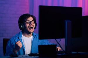 A young man, wearing glasses and headphones, is sitting in front of a computer screen in a dimly lit room. He is smiling and laughing, with his fist raised in the air, as he plays a game. photo