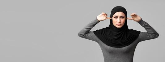 Hear no evil. Arabic girl in hijab closing ears with fingers and looking at camera with angry face expression, gray background with free space photo