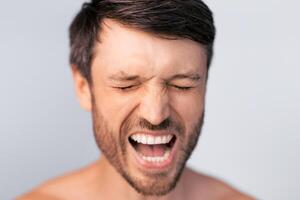 A close-up portrait of a man with dark hair and a beard, shouting with his eyes closed, captured against a light gray background. photo