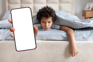 African American child lies in bed under the covers, displaying frustration while looking at a smartphone held out in front of them. The setting is a cozy bedroom with simple decor. photo