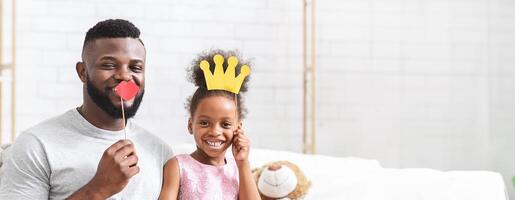A joyful black father and his daughter are sharing a lighthearted moment, both smiling and posing for fun with playful props. The father holds a red heart-shaped object, while the girl wears a crown photo
