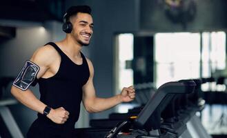 Handsome Young Muscular Arab Man In Wireless Headphones Jogging On Treadmill At Gym, Sporty Motivated Middle Eastern Guy Listening Music While Running, Enjoying Making Sports, Copy Space photo