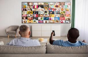 Two men sit on a couch in a living room, facing a large television screen displaying a grid of various images. The man on the right is holding a remote control in his hand photo