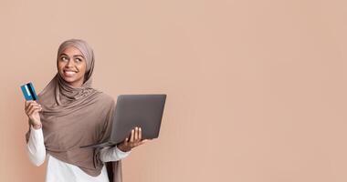 Online payments concept. Thoughtful black islamic girl in headscarf with laptop and credit card and looking away, yellow background with empty space photo