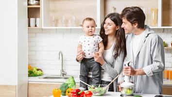 A happy family is preparing a healthy meal together in their modern kitchen. The mother is holding their baby, who is looking at the camera with a curious expression, copy space photo