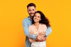 A man with a beard is embracing a woman with long dark hair. They are both smiling and looking directly at the camera. They are standing in front of a solid yellow background. photo