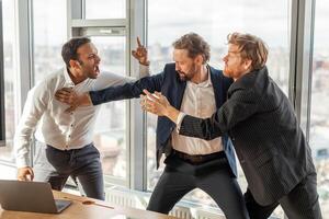 Three men in suits are engaged in a heated argument in a modern office setting. The man in the white shirt is yelling at the man in the blue suit, who is pushing him away photo