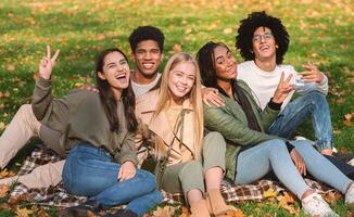 Multiethnic Group of Happy Teenagers Having Fun In Autumn Park, sitting on the ground and posing at camera photo
