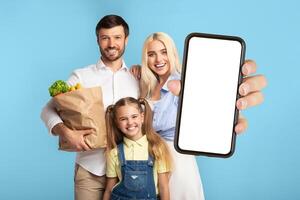 A happy family, father, mother, and daughter, stand together in front of blue background. Father holds brown paper bag filled with groceries, while the mother holds a smartphone with a blank screen photo