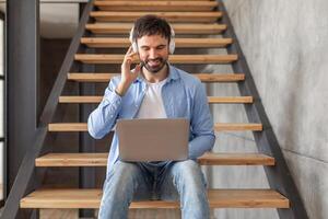 A man sits on a staircase with a laptop in his lap. He is wearing headphones and adjusting them while looking at the screen. He is wearing a blue button down shirt and jeans. photo