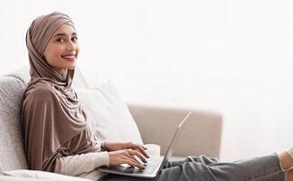 Online Degrees Concept. Smiling Muslim Girl In Hijab Using Laptop At Home, Studying In Internet, Learning Foreign Languages, Side View, Copy Space photo