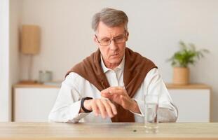 Elderly Gentleman Checking Time With Smartwatch Sitting At Table With A Pill And Glass Of Water At Home photo
