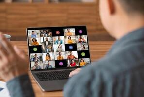 A man sits at a desk, focused on his laptop screen, participating in a large online conference meeting with a diverse group of people. He is engaged in the discussion, with his hand raised photo