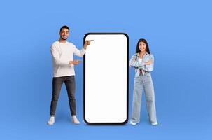 A man and a woman are standing side by side, both pointing excitedly at a blank screen in front of them. The background is a solid blue color. photo