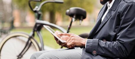 A businessman is sitting on a bench in a park, using a digital tablet with his bicycle close by. The atmosphere is tranquil and efficient, showing a modern work-life balance, cropped photo