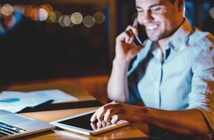Business Application. Businessman Using Digital Tablet Talking On Phone Working Late At Night In Modern Office. Selective Focus, Low Light photo