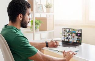 A man sitting at his desk in a home office during the daytime, actively participating in a virtual business meeting on his laptop while taking notes in a notebook. photo