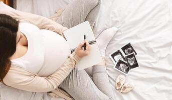 Pregnant woman making packing list for maternity hospital, preparing for labor at home, top view, empty space photo