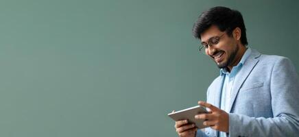 Indian man is engaged with a tablet, smiling warmly in a contemporary indoor space. He wears a light suit and glasses, creating a relaxed and professional atmosphere. photo