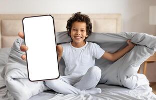 African American young boy with curly hair smiles brightly while holding a smartphone. He is wrapped in a blanket, seated on a neatly made bed in a warm, inviting bedroom setting. photo
