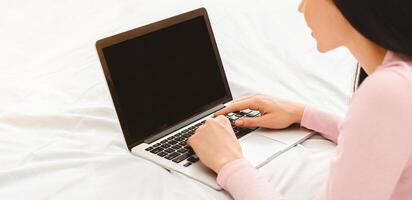 A woman is seated on a bed, attentively using a laptop computer with blank screen in front of her. She appears focused on her screen, typing and clicking, cropped photo