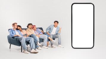 A group of diverse friends, dressed casually in jeans and t-shirts, sit on a blue couch and look at a giant smartphone. One friend points at the blank screen. photo