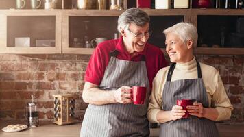A senior couple wearing aprons stands together in a kitchen, each holding a red mug of coffee. They are smiling and looking at each other, enjoying a moment of connection and togetherness. photo