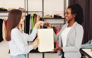 Two multiracial women are browsing through racks of clothes in a busy clothing store. They are holding clothing items and discussing their choices photo