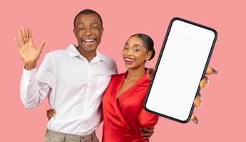 Happy black couple posing in front of a pink background. The woman is wearing a red dress and holding a smartphone with a blank screen, while the man is wearing a white shirt and waving photo