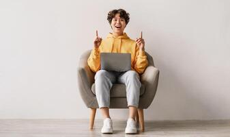 Millennial Asian man having creative idea, gesturing eureka with both hands, pointing up, offering space for design, sitting in armchair with laptop pc against white studio wall, copy space photo