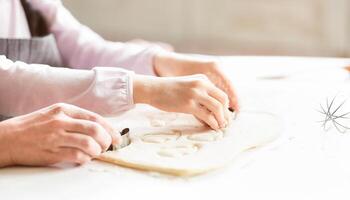 A close-up photo of a woman and a child cutting out shapes from dough using cookie cutters. They are working together in a kitchen environment. The woman is on the left, the child is on the right.
