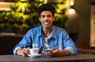 A young black guy is seated at a table, where he is observed with a cup of coffee in front of him. He appears to be enjoying a moment of relaxation while sipping his beverage. photo