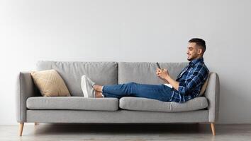 Cool Gadget And Application. Portrait of young smiling Arab man holding mobile phone, typing sms message, sitting on the sofa in living room. Guy browsing internet, surfing web, using app, free space photo