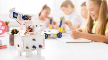 A close-up shot of a white robotic arm on a table in a classroom setting. The robot is in focus and there are blurred children in the background who are building or working on projects. photo