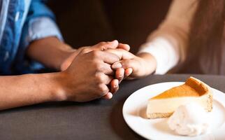 Two individuals are pictured holding hands at a table in a cozy cafe. A slice of cheesecake on a plate with a dollop of whipped cream is in front of them, suggesting a warm and intimate moment photo