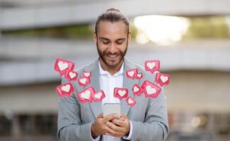 A young man with a beard and a bun hairstyle is looking at his smartphone while smiling. He is wearing a gray suit jacket, and several digital hearts representing social media likes photo