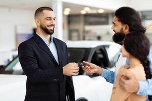 A well-dressed salesman is standing in a car showroom, presenting car keys to a cheerful Indian couple, symbolizing the finalization of a vehicle purchase photo