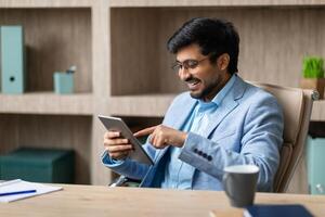 A smiling Indian businessman sits at a desk in his office, looking at a tablet and using his finger to navigate the screen. There is a cup of coffee on the desk and a notebook with a pen beside it. photo