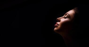 A close up shot of a womans face with eyes closed, lit from the side, with a dark background. She is turned slightly away from the camera. Her lips are closed and she appears to be relaxed. photo