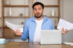 A man with a surprised expression is seated at a desk in a home office, holding papers in his hands. A laptop is open in front of him, suggesting a moment of unexpected news or work-related shock. photo