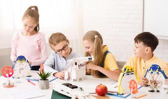 Four children, three boys and one girl, are working on building robots in a classroom setting. They are all focused on their tasks and seem to be enjoying themselves. The classroom is bright photo