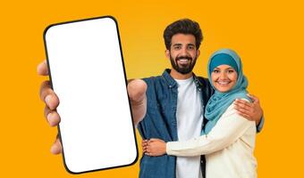 A young muslim eastern man and woman, both smiling, stand together against a bright yellow background. The man holds out a smartphone with a blank screen towards the viewer. photo