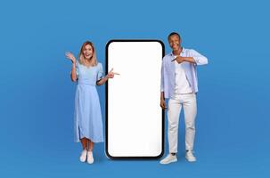 A man and woman are pointing at a blank screen against a solid blue background. Both individuals are gesturing towards the screen, displaying engagement and interaction. photo